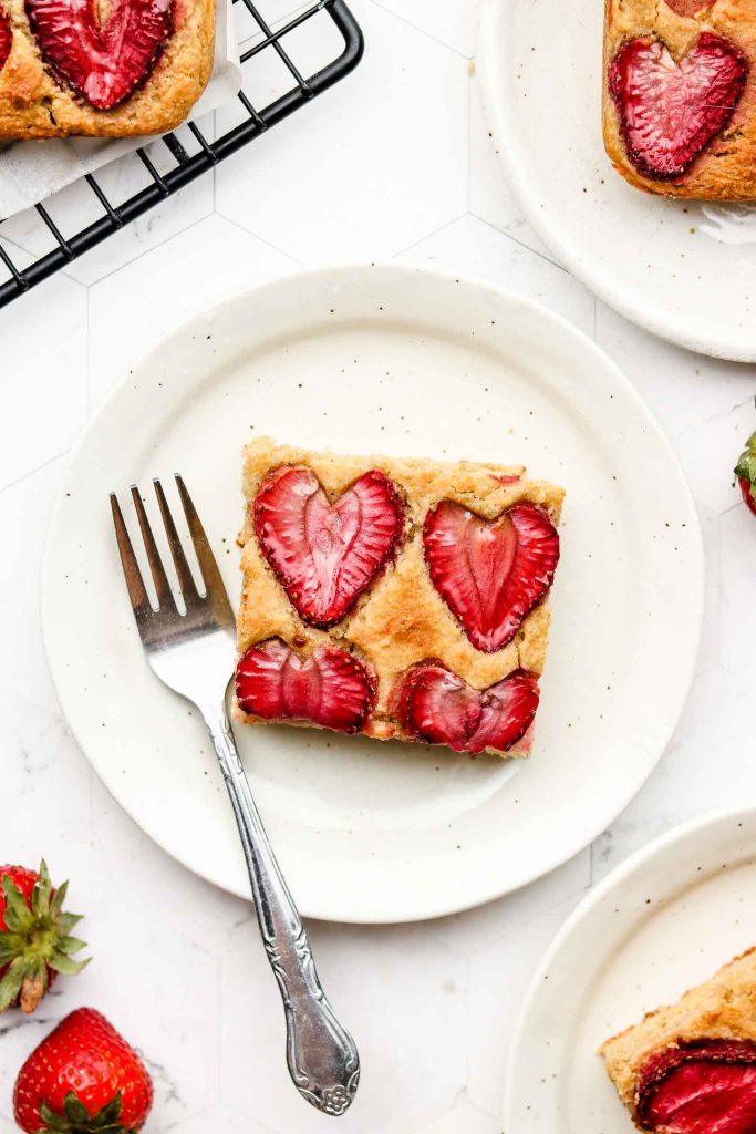 square piece of lemon sheet cake with strawberries on top on a white speckled plate and a fork on the side 