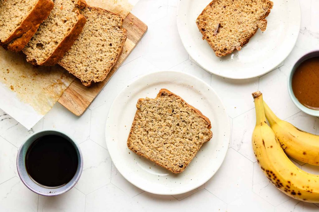 slices of healthy banana bread on white speckled plates with coffee on the left side and bananas on the right side