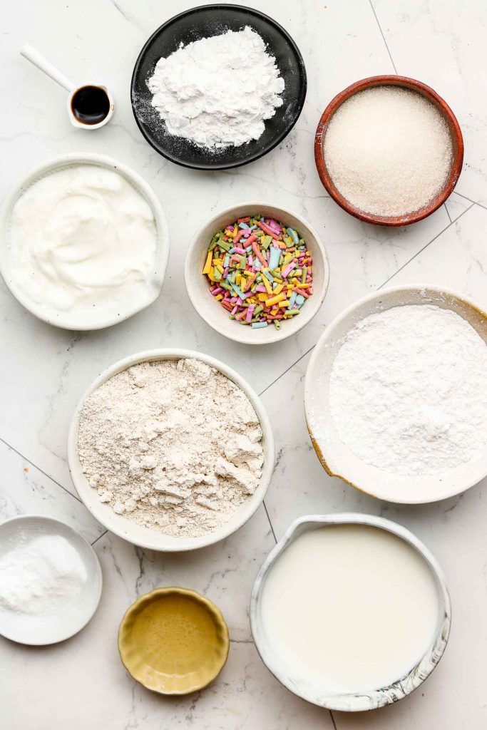 ingredients for vegan funfetti cake in bowls on a marble back drop