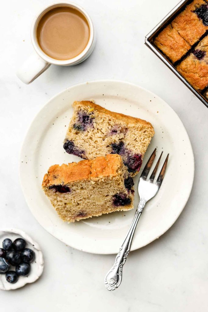 two slices of lemon blueberry loaf on a plate with a fork and coffee on the top left corner