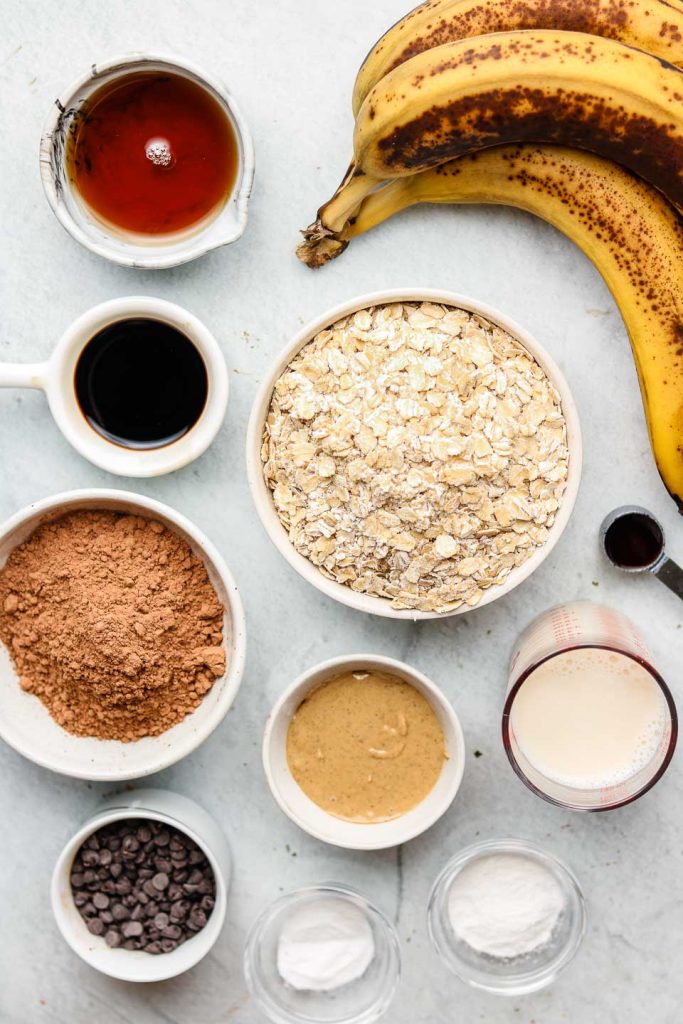 ingredients for healthy chocolate banana muffins in white bowls on a blue backdrop