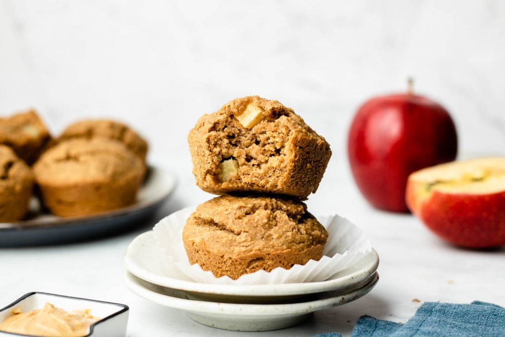 healthy oat apple muffins stacked on a speckled small plate with a bite taken out of the top muffins and apples in the background